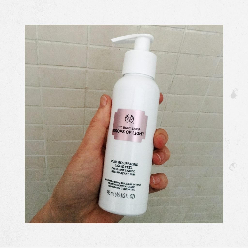 hand holding The Body Shop's Drops of Light Pure Resurfacing Liquid Peel , backdrop is a shower 