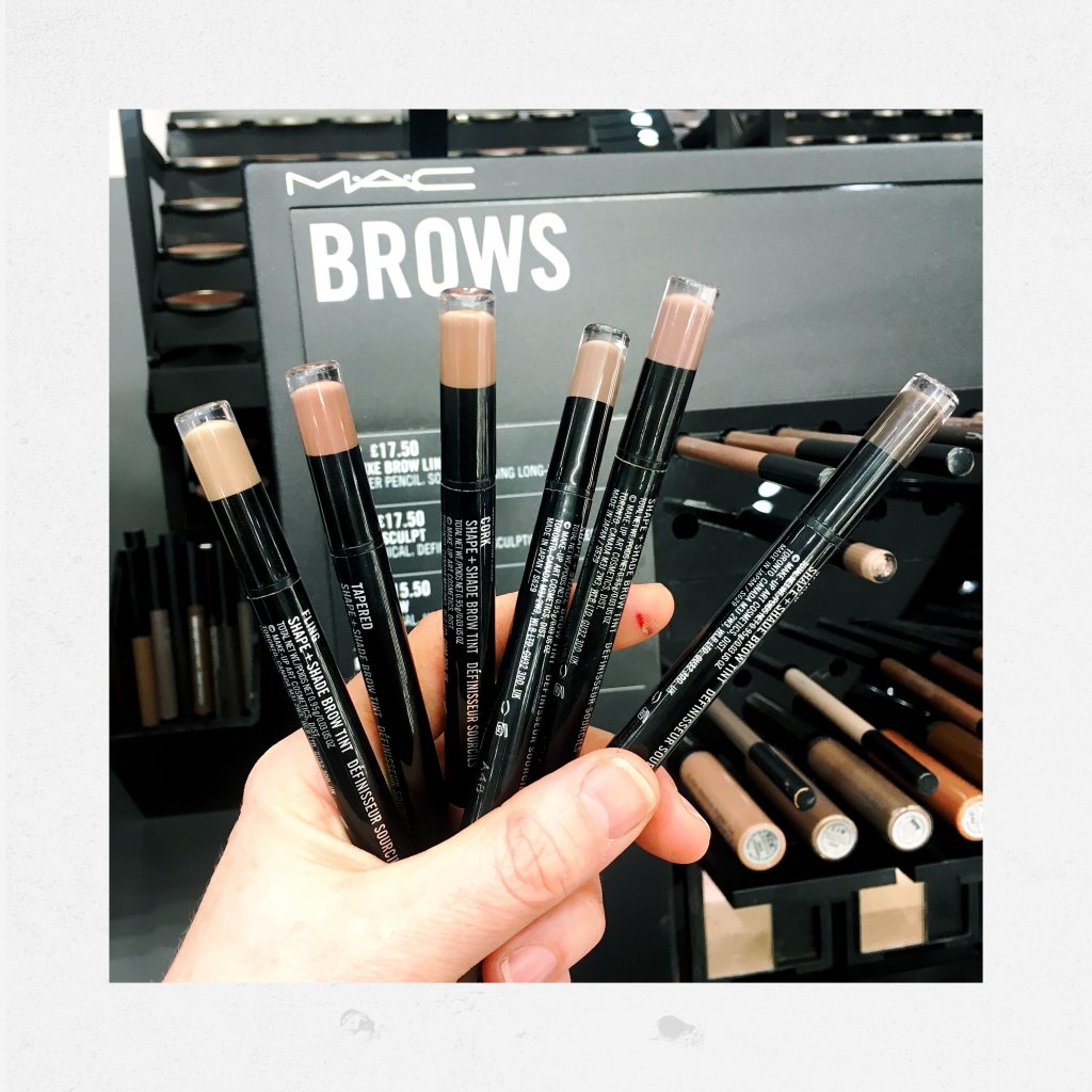 Hand holding 6 shades of MAC Shape and Shade Brow Tint which is in pen form. the sign behind reads "Brows" 